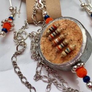 May include: A silver necklace and earrings set with a cork pendant featuring orange and blue beads. The pendant is decorated with silver-colored resistors. The tag reads "King Blaz Creations".