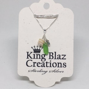 May include: A silver necklace with a green glass charm, white and pink shell charms, and a silver crown charm. The necklace is on a white card with the text "King Blaz Creations Sterling Silver".