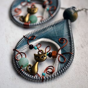 Puede incluir: Un par de pendientes de alambre envueltos en color azul verdoso y blanco con un dije de gato, cuentas turquesas y cuentas de madera.