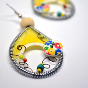 Puede incluir: Un par de pendientes amarillos en forma de lágrima con un marco de alambre plateado y un botón floral colorido. Los pendientes están hechos con un hilo amarillo y blanco y tienen una cuenta de madera en la parte superior.
