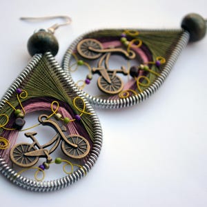 Puede incluir: Un par de pendientes colgantes con un marco de alambre plateado y un diseño de hilo rosa, verde y marrón. Un pequeño amuleto de bicicleta de metal se encuentra en el centro de cada pendiente.