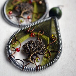 Puede incluir: Un par de pendientes verdes y plateados con un diseño de árbol y detalles de alambre de cobre. Los pendientes están hechos de alambre envuelto alrededor de hilo verde y tienen un pequeño amuleto de árbol en el centro.