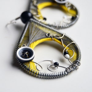 Puede incluir: Un par de pendientes hechos con alambre, hilo amarillo y gris, y cuentas negras y blancas. Los pendientes tienen un diseño de botón con un botón negro en el centro.