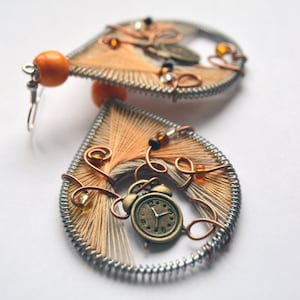 Puede incluir: Un par de pendientes hechos a mano con forma de lágrima. Los pendientes están hechos con alambre de cobre, cuentas y un pequeño dije de reloj. Los pendientes son de color marrón claro con cuentas naranjas.