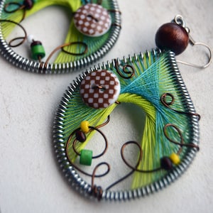 Puede incluir: Un par de pendientes hechos a mano con forma de lágrima. Los pendientes están hechos con alambre de plata, hilo verde y azul, cuentas de madera marrón y botones blancos con lunares marrones.