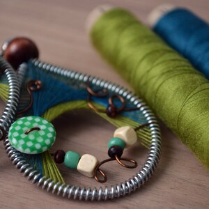 May include: A pair of handmade earrings with green and blue thread wrapped around a metal frame. The earrings feature green and white checkered buttons and brown and white beads.