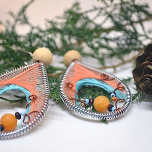 May include: Handmade teardrop earrings with orange, teal, and copper thread detailing. The earrings have a silver metal frame, wooden bead tops, and small decorative beads. The background includes greenery and a pinecone.