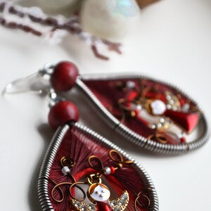 May include: A pair of red and silver dangle earrings featuring a small angel charm. The earrings are crafted from wire and red thread.