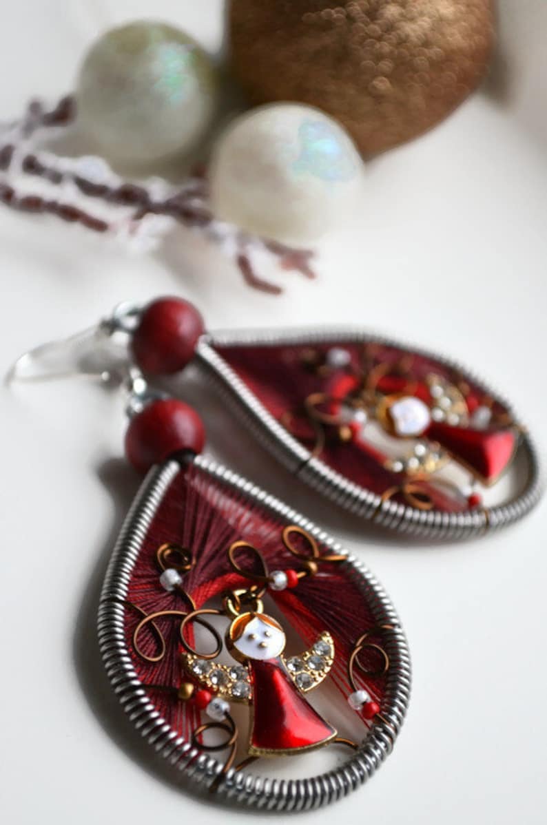May include: A pair of red and silver dangle earrings featuring a small angel charm. The earrings are crafted from wire and red thread.