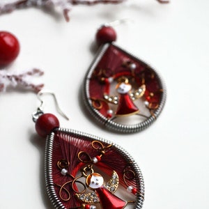 May include: A pair of red and silver dangle earrings with a small angel charm in the centre. The earrings are made with red thread wrapped around a silver wire.