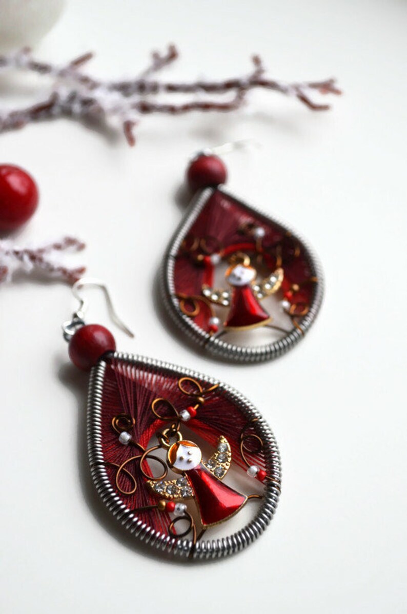 May include: A pair of red and silver dangle earrings with a small angel charm in the centre. The earrings are made with red thread wrapped around a silver wire.