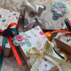 May include: Five small gift boxes wrapped in festive paper with colorful ribbons and bows. The boxes are decorated with various holiday themes, including reindeer, Christmas trees, and snowflakes.