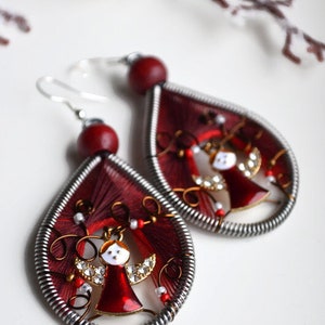 May include: A pair of red and silver dangle earrings with a small angel charm in the centre. The angel is wearing a red dress and has white wings.