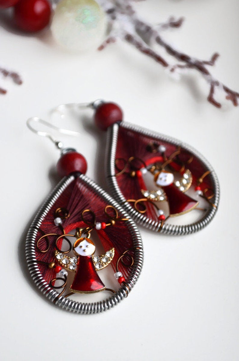 May include: A pair of red and silver dangle earrings with a small angel charm in the centre. The angel is wearing a red dress and has white wings.
