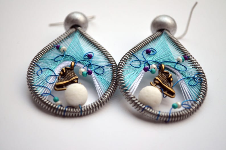 May include: A pair of teardrop-shaped earrings with a silver wire frame and blue and white thread woven into a design. Each earring features a small white bead and a bronze-colored ice skate charm.