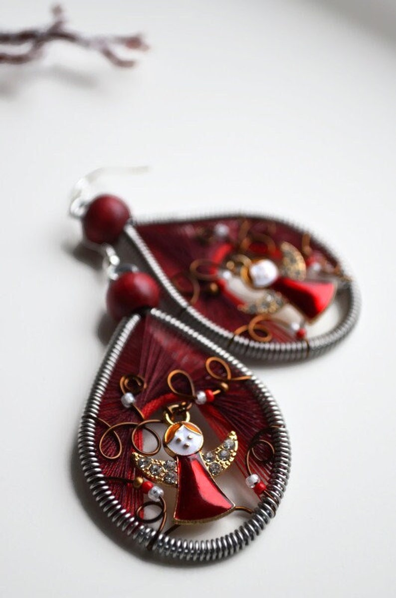 May include: Red and silver teardrop-shaped earrings with a small red angel charm in the centre. The earrings are made with wire and red thread.