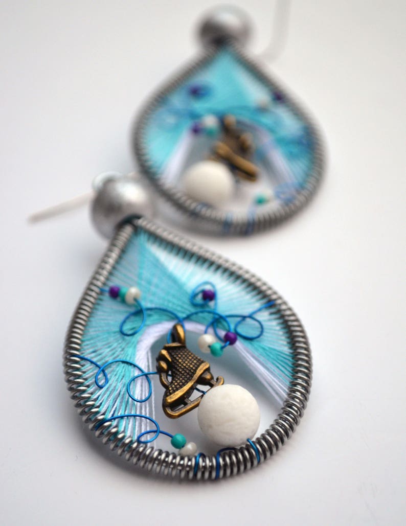 May include: A pair of teardrop-shaped earrings made of wire wrapped with blue, white, and turquoise thread. Each earring features a small, white bead and a bronze-colored charm of a figure ice skating.