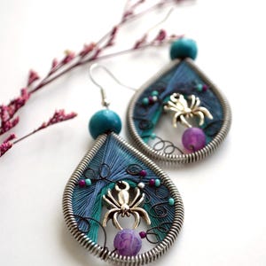 May include: Teardrop-shaped earrings with a silver wire frame, featuring blue and teal threadwork. Each earring has a silver spider charm, purple and teal beads, and a turquoise bead at the top. A sprig of pink flowers is in the background.