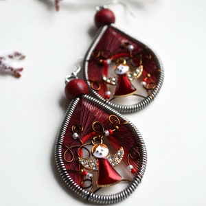 May include: A pair of red teardrop-shaped earrings with a silver wire frame. Each earring features a small gold angel with red wings and a white face. The angel is surrounded by red and white beads.