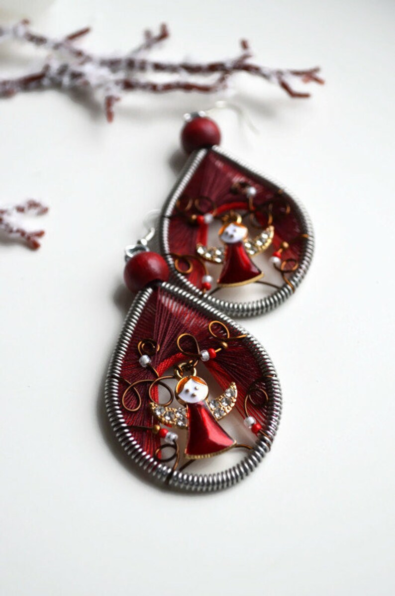 May include: A pair of red teardrop-shaped earrings with a silver wire frame. Each earring features a small gold angel with red wings and a white face. The angel is surrounded by red and white beads.