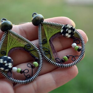 Puede incluir: Un par de pendientes verdes y marrones con alambre envuelto alrededor de un hilo verde y marrón. Los pendientes tienen un botón blanco y negro y cuentas verdes y marrones.