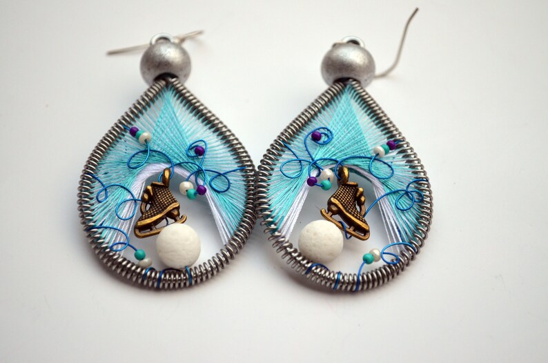 May include: A pair of teardrop-shaped earrings with a winter theme. Each earring features a white and blue wire wrapped design with a small white bead, a bronze ice skate charm, and a silver hook.