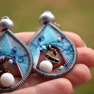 May include: A pair of dangle earrings with a blue, white, and purple thread design. Each earring features a small white bead and a silver charm shaped like an ice skate.
