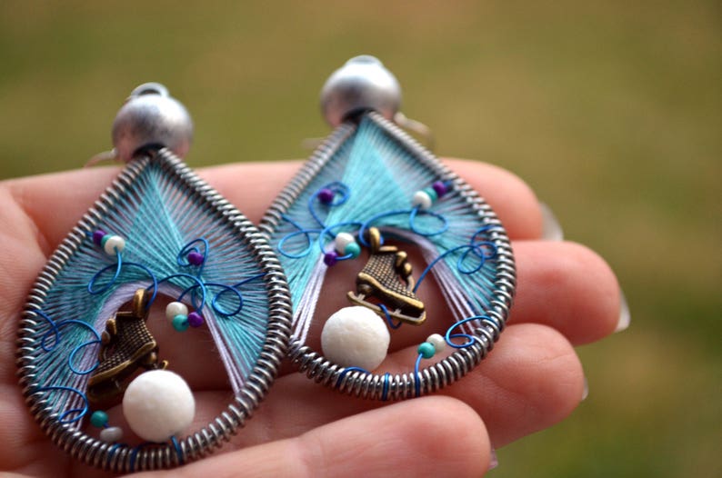 May include: A pair of dangle earrings with a blue, white, and purple thread design. Each earring features a small white bead and a silver charm shaped like an ice skate.