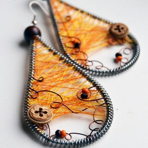 Puede incluir: Un par de pendientes de color naranja y marrón en forma de lágrima con un diseño de alambre y hilo. Los pendientes tienen un botón y cuentas.