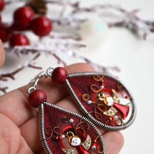 May include: A pair of red and silver dangle earrings featuring a small angel charm. The earrings are crafted from wire and red thread.