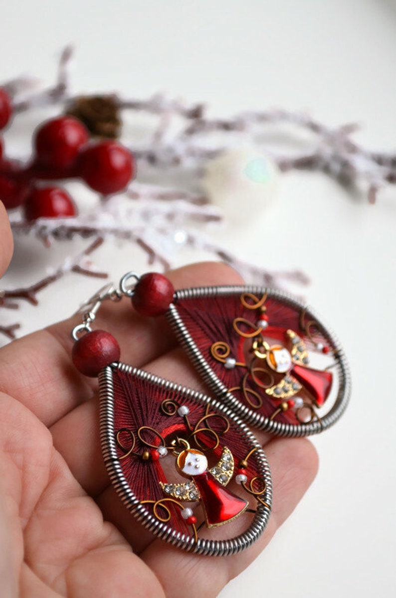 May include: A pair of red and silver dangle earrings featuring a small angel charm. The earrings are crafted from wire and red thread.