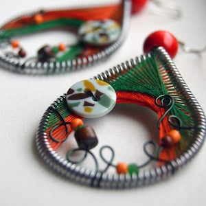 Puede incluir: Un par de pendientes hechos a mano con un esquema de color verde, rojo y naranja. Los pendientes están hechos con alambre, cuentas y botones. Los botones son de un patrón de camuflaje blanco y marrón.