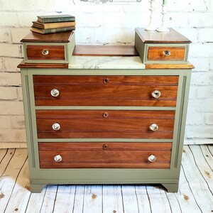 May include: A vintage dresser with three drawers, painted in a sage green color with a wood top. The dresser has crystal knobs and a marble top. The dresser is labeled "Reimagined by Elizabeth Anne Est. 2020".