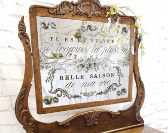 May include: A vintage wooden vanity with a mirror. The mirror has decorative floral accents and French text that reads "TU ES ET TUSER toujours la plus BELLE SAISON de ma vie." The vanity is brown with ornate carvings and drawer pulls.
