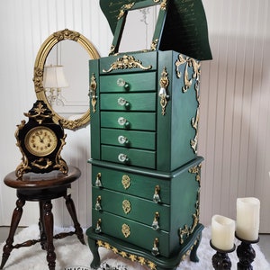 May include: A green jewelry armoire with gold accents and crystal drawer pulls. The armoire has a mirrored top with a lift-up lid. The armoire is sitting on a white fluffy rug. The text "RE/MAGINED BY ELIZABETH ANNE EST. 2020" is on the bottom of the armoire.