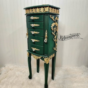 May include: A tall, dark green jewelry armoire with six drawers and ornate gold accents. The armoire has a decorative top and bottom, with crystal-like drawer pulls. The legs are curved and painted green. The armoire is approximately 60 inches tall.