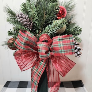 May include: A festive Christmas decoration featuring faux evergreen branches, red and gold ornaments, and pinecones. A large plaid bow with red and green accents adds a decorative touch. The decoration is mounted on a green stake.