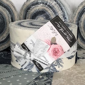 Peut inclure: Trois rouleaux de tissu enroulés dans du plastique transparent, présentant un motif en spirale de bandes de tissu crème, gris et bleu foncé. Un rouleau central porte une étiquette en papier blanc avec le texte Moda Fabrics Supplies et une rose.