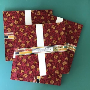 May include: Three stacks of square fabric pieces bundled with white paper bands. The fabric features a dark red background with a repeating pattern of small yellow floral clusters. A label on the center stack reads Henry Glass & Co. Inc. 10" x 10".