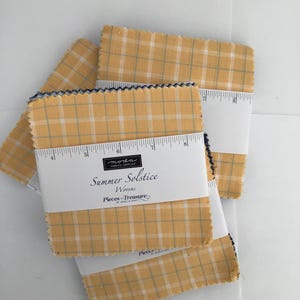 May include: A stack of yellow and white plaid fabric squares with a dark blue zigzag edge. The fabric squares are wrapped with a white label that reads "Summer Solstice Wovens" and "Pieces of Treasure".