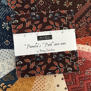 May include: A collection of fabric squares in various patterns and colors, including floral, paisley, and geometric designs. The dominant colors are burgundy, navy, and cream. A white label with the text "Pamelia's Path 1850-1880 by Betsy Chutchian" is placed on top of the fabric.