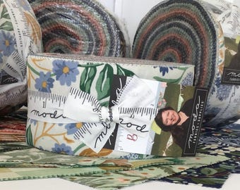 May include: Fabric rolls with floral and leaf patterns in shades of blue, green, and cream. The fabric is wrapped with a white ribbon printed with ruler markings and the word "moda". A business card with the "moda fabrics-supplies" logo is attached.