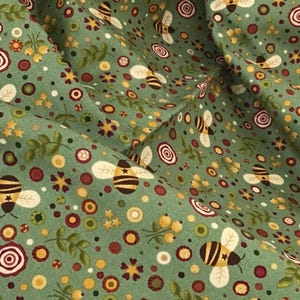 May include: Close-up of a green fabric with a repeating pattern of bees, flowers, and circular designs in shades of yellow, brown, and red. The fabric has a slightly wrinkled texture, suggesting it is a soft material.