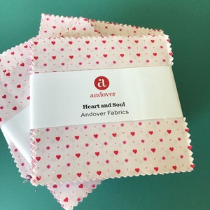 May include: A stack of pink fabric squares with a white background and a red heart pattern. The fabric squares have a scalloped edge. The top square has a white label with the Andover logo and the text "Heart and Soul Andover Fabrics".