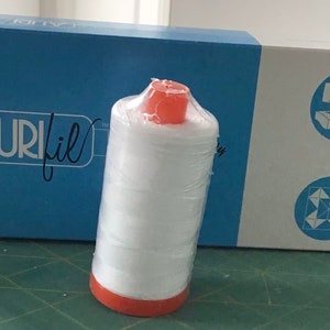 May include: A white spool of Aurifil thread with an orange plastic top and a blue and white box with the Aurifil logo. The box also has a sewing machine and a quilt block design.