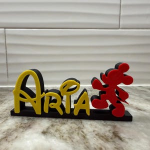 May include: A black base with the name "Aria" in yellow and black lettering, and a red and black Mickey Mouse figure. The name is in a playful font, and the Mickey Mouse figure is in a running pose.