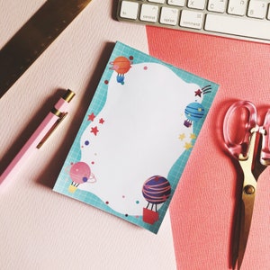 May include: A blue and white notepad with a whimsical space theme. The notepad features a white background with a blue border and a wavy design. There are colourful planets and stars on the notepad. The notepad is on a pink surface with a gold ruler and a pink pen.