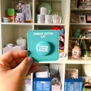 May include: A turquoise blue pinback button with white text that reads "taking a deep breath" on a white background. The button is 1.25 inches in diameter and is displayed on a turquoise blue card with the words "Pinback Button 1.25" printed in white.