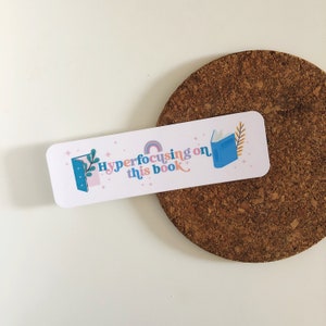 May include: White bookmark with a blue book, rainbow, and stars. The text reads "Hyperfocusing on this book."