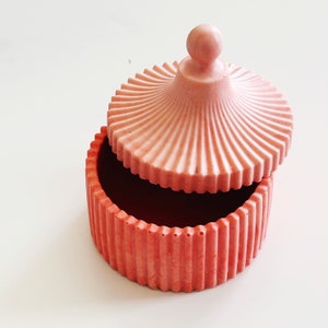 Könnte beinhalten: Eine rosafarbene Keramik-Schmuckschatulle mit geripptem Design und einem gewölbten Deckel. Der Deckel hat ein geriffeltes Design und einen kleinen Knopf oben.